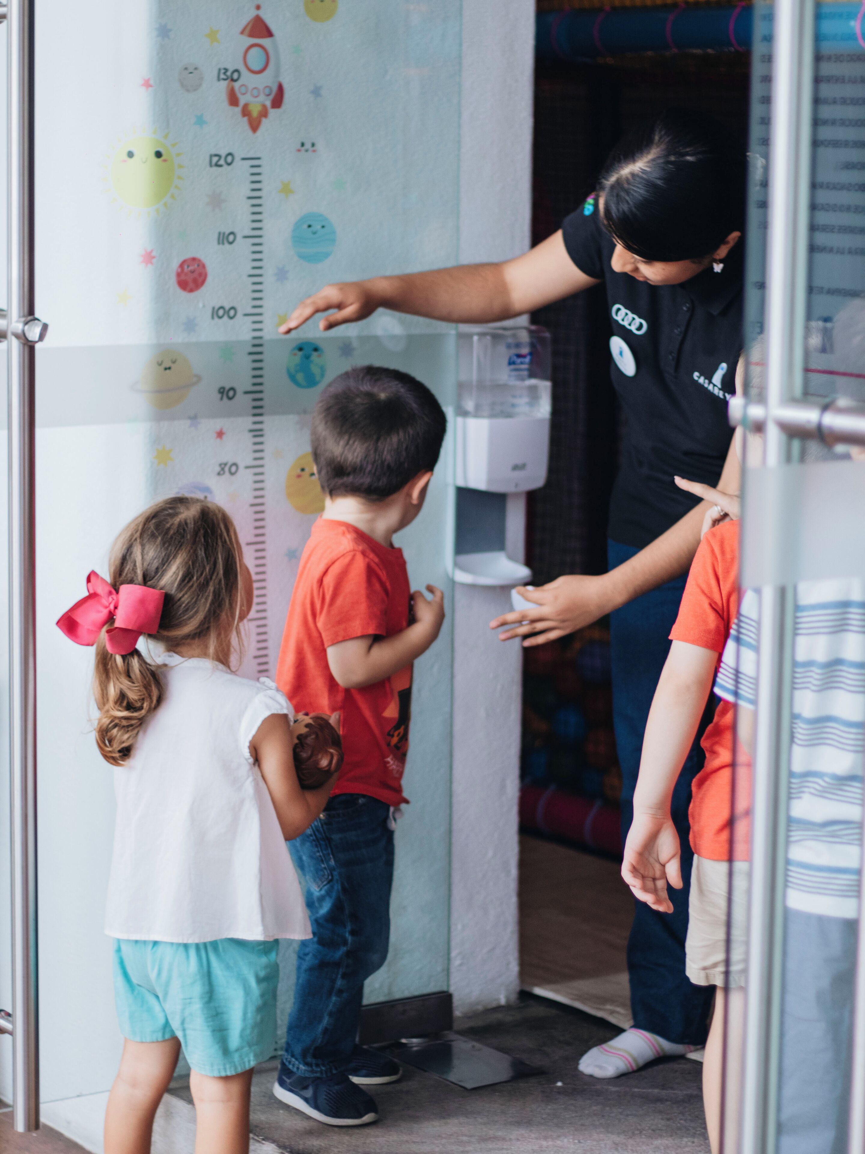 Un niño y un adulto interactúan cerca de un dispensador de agua en una habitación luminosa y decorada, con una tabla de medidas en la pared.