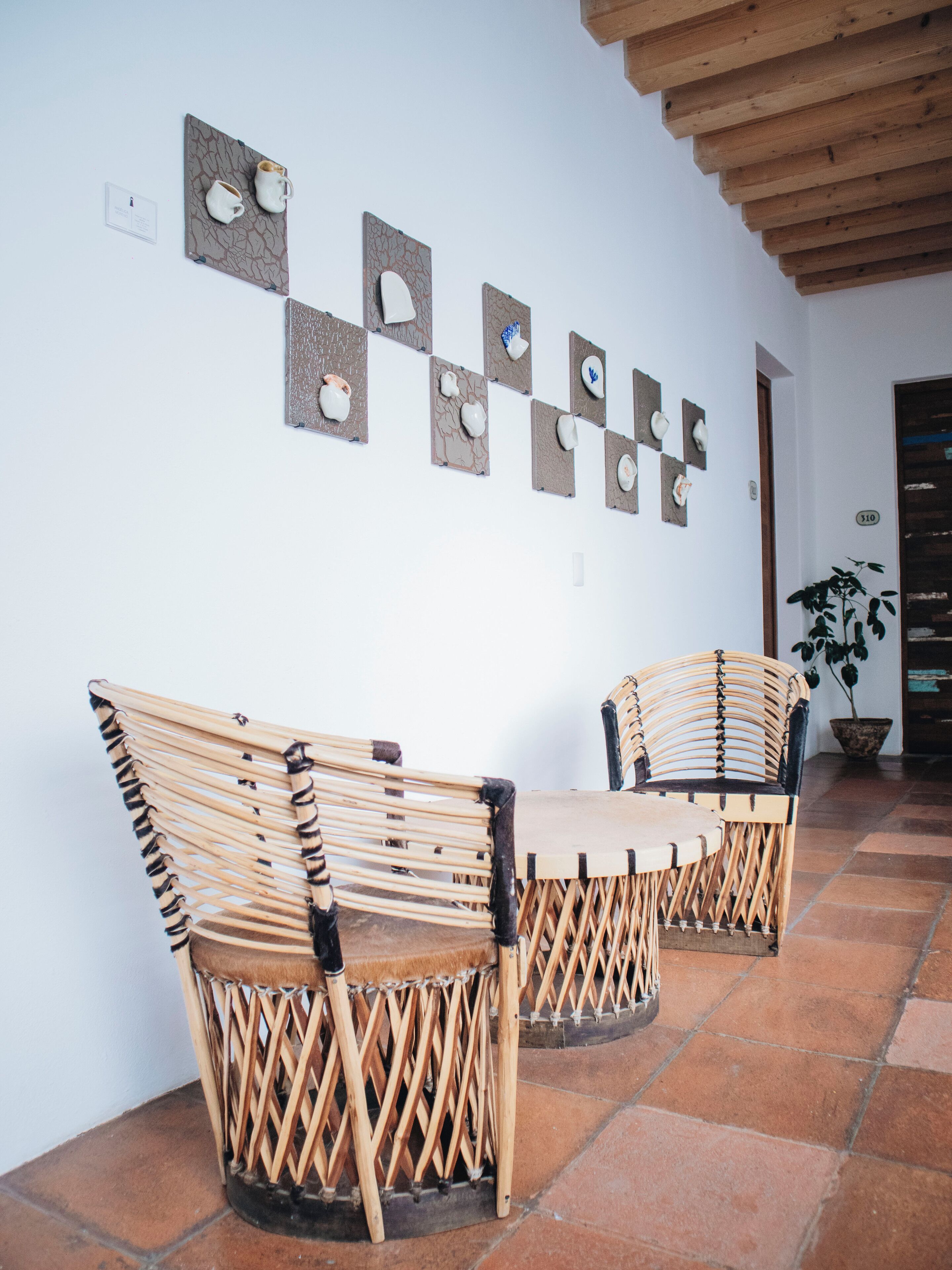 Two wicker chairs with a woven backrest and seat, positioned in front of a wall with a series of square frames, each containing a white object.