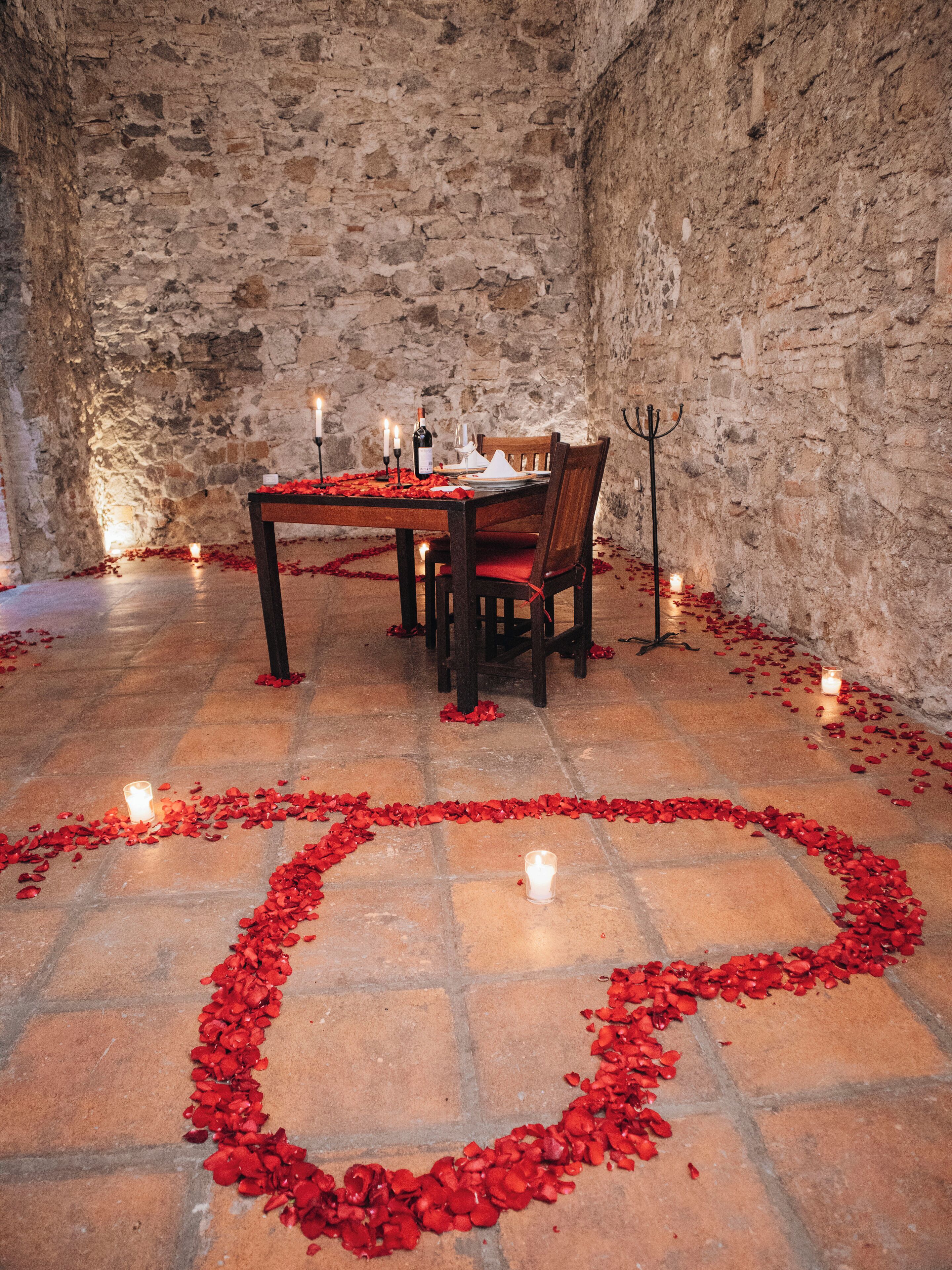 Una habitación con paredes de piedra, con una mesa lista para dos personas, rodeada de pétalos de rosa roja formando un corazón en el suelo.