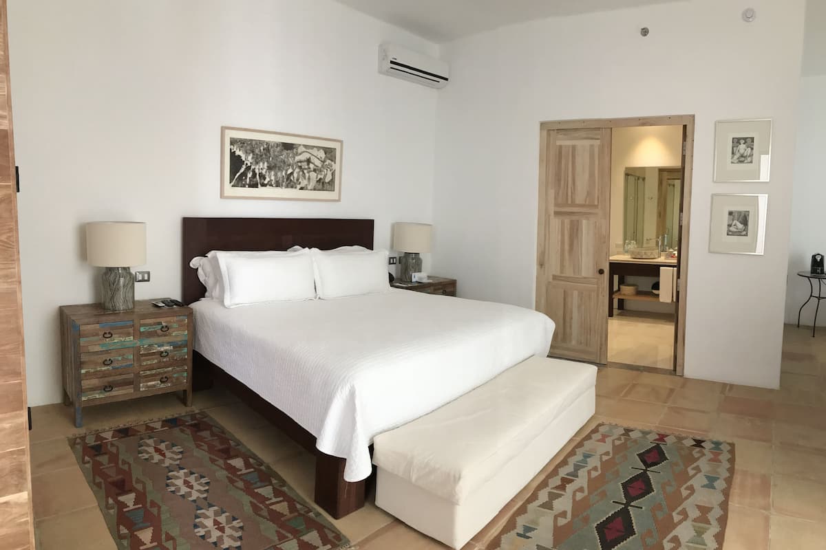 Un dormitorio ordenado con una cama grande, mesitas de noche con lámparas, un cabecero de madera y una puerta que da al baño.