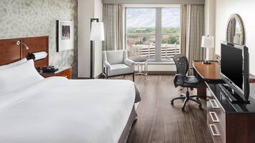 A hotel room with a large bed, a desk with a TV, a chair, a desk lamp, and a view of the city through the window.