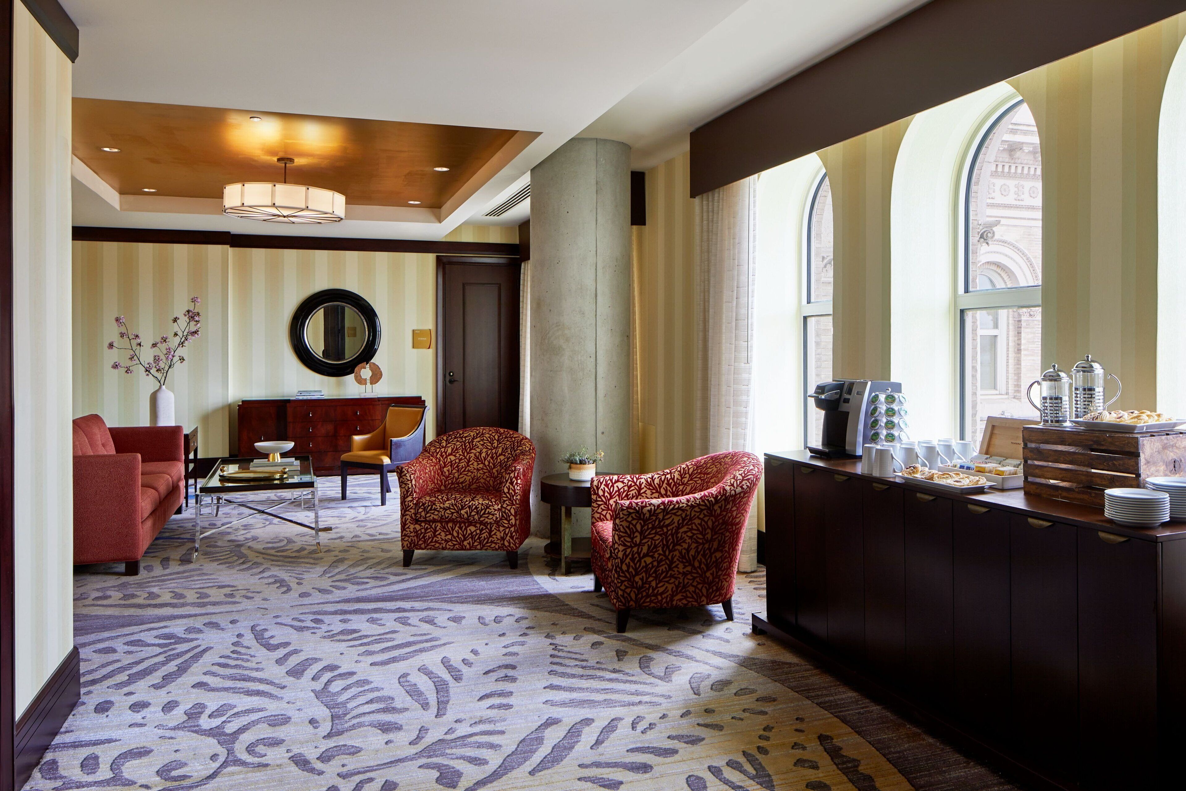 Eine Hotelhalle mit einem gemusterten Teppich, roten Sofas, gemusterten Sesseln, einer Kaffeestation und großen Fenstern mit Rundbögen.