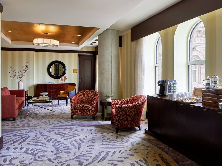 Eine Hotelhalle mit einem gemusterten Teppich, roten Sofas, gemusterten Sesseln, einer Kaffeestation und großen Fenstern mit Rundbögen.