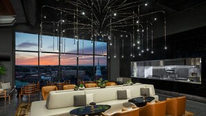 Un área de descanso moderna con un sofá, sillas y una mesa de comedor. Amplias ventanas ofrecen vistas del paisaje urbano y una puesta de sol.