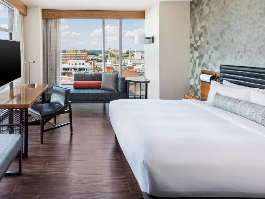 A hotel room with a large bed, a desk with a flat-screen TV, a sofa, and a view of the cityscape.