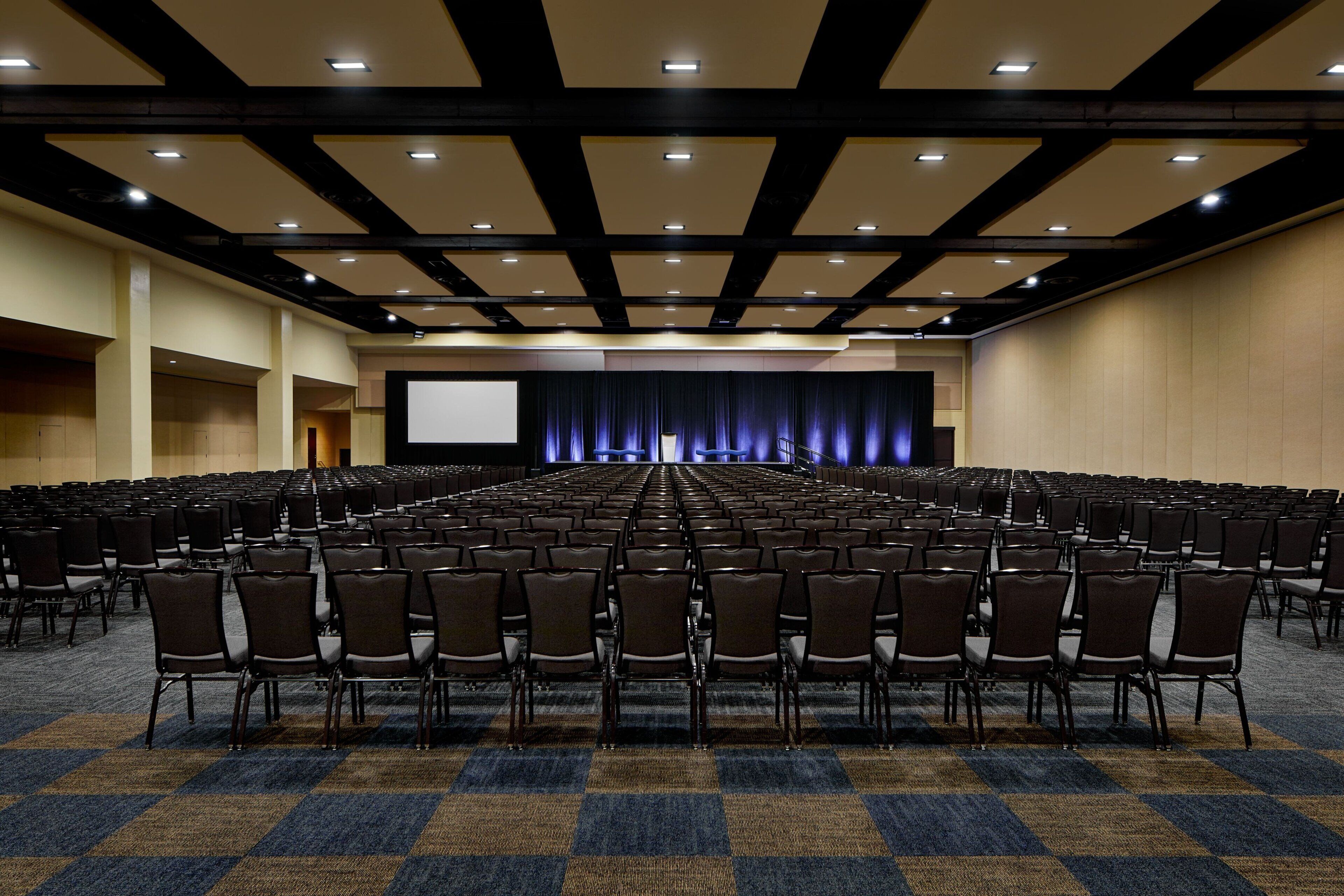 A large conference room with rows of chairs, a stage with a screen, and a carpeted floor.