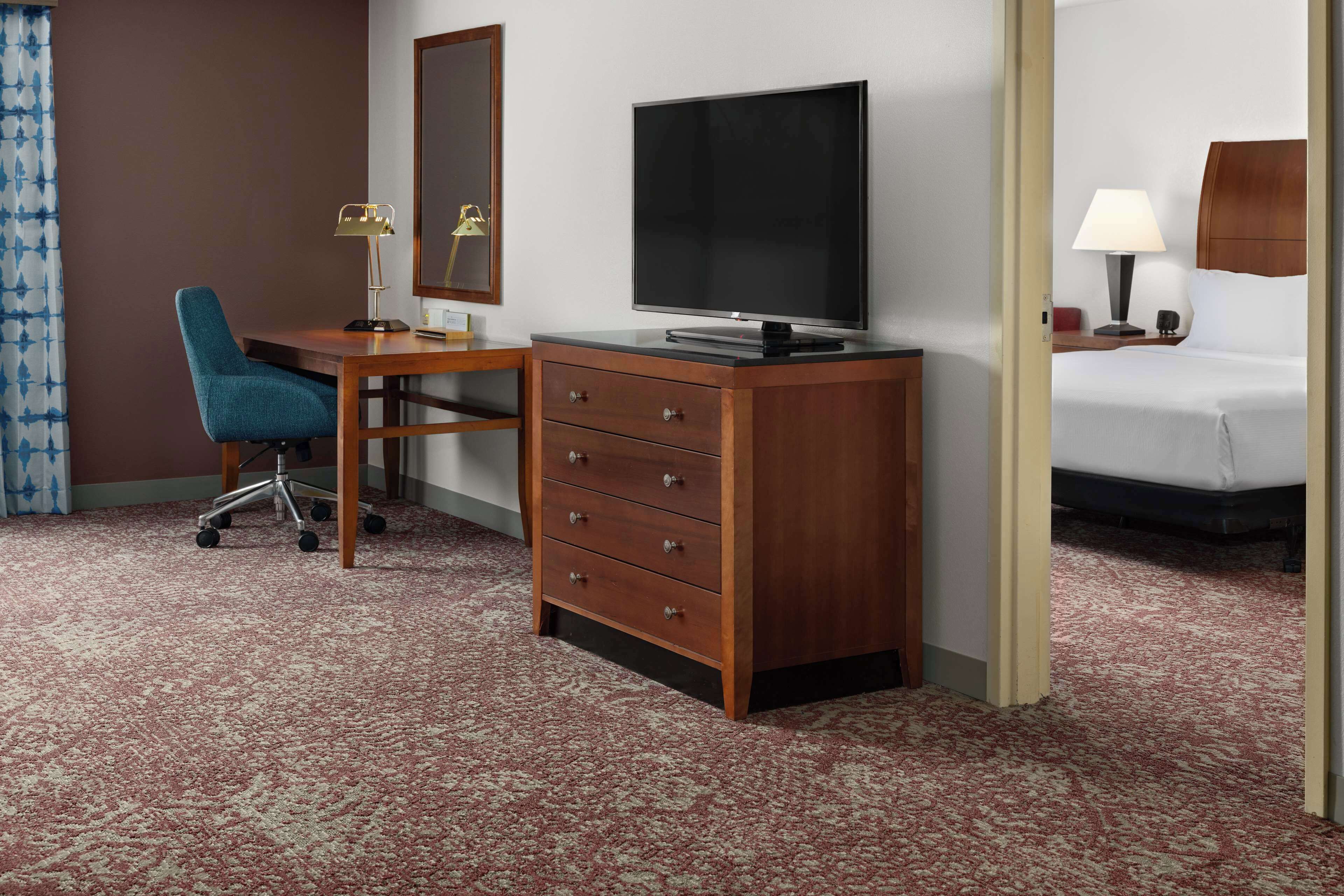 A hotel room with a wooden desk, a television, a bed, and a nightstand with a lamp.