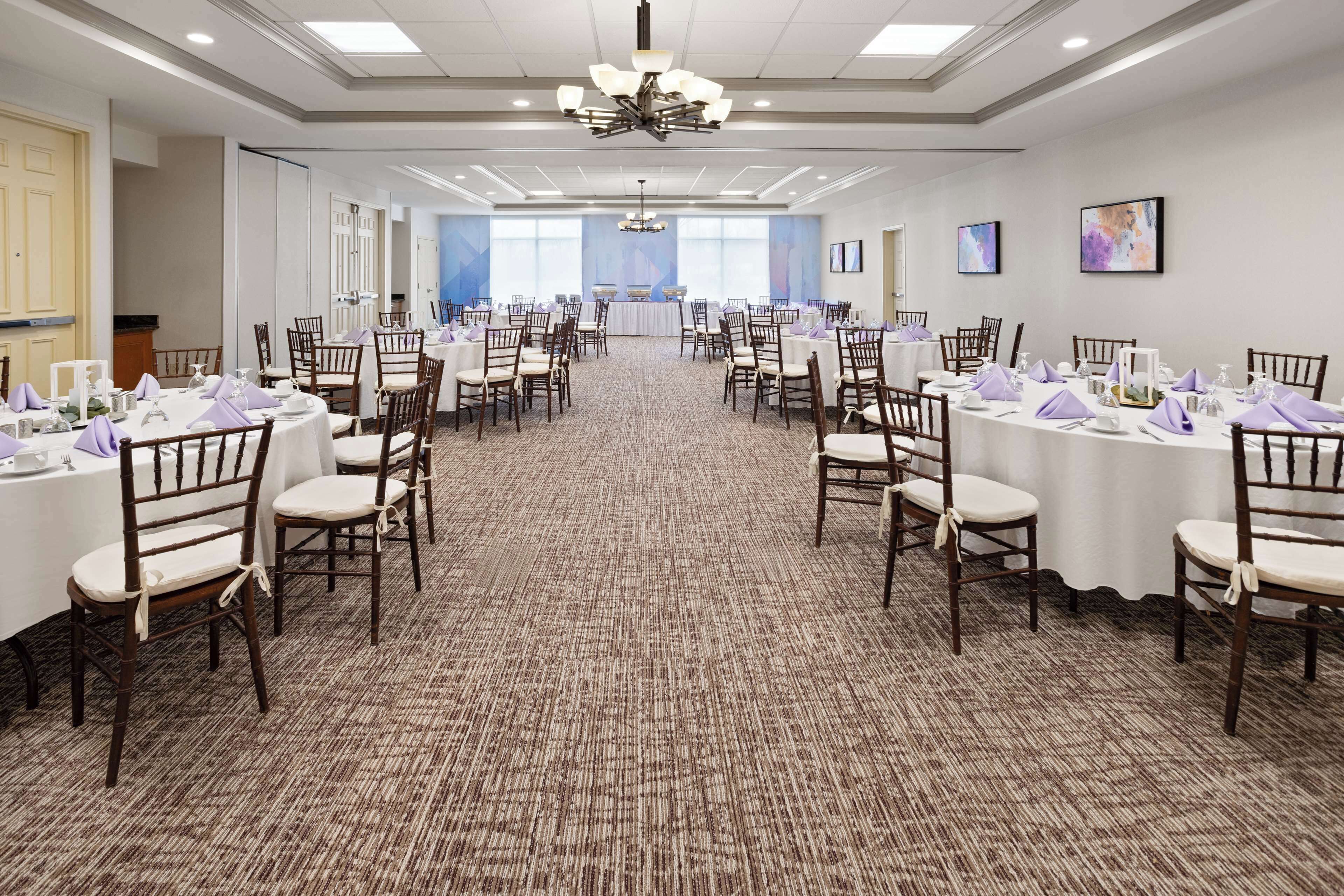 A large conference room with multiple round tables, each set with white tablecloths, chairs, and purple napkins.