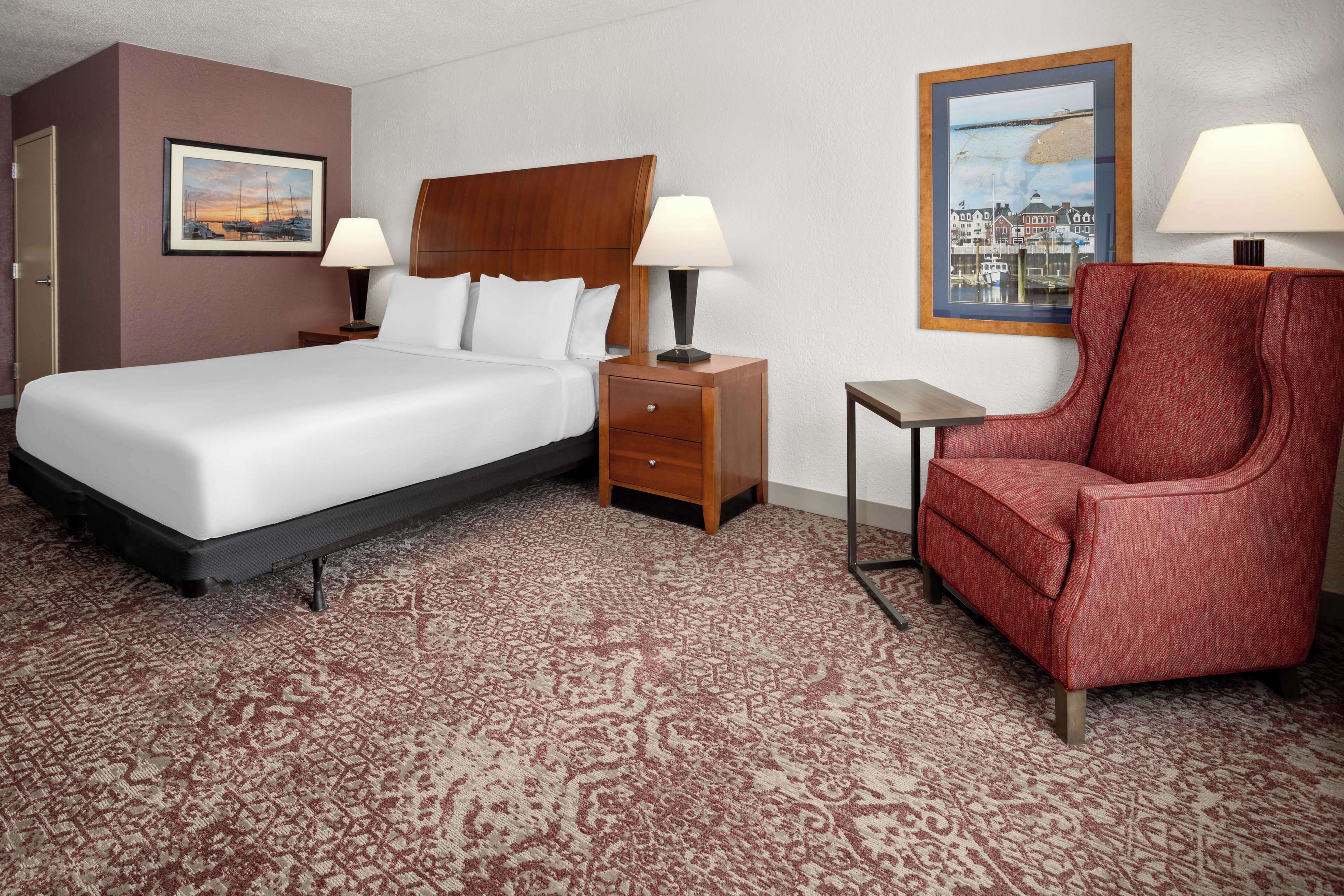 Habitación de hotel con cama, dos mesitas de noche con lámparas, un sillón rojo, una pequeña mesita auxiliar y dos cuadros en las paredes.