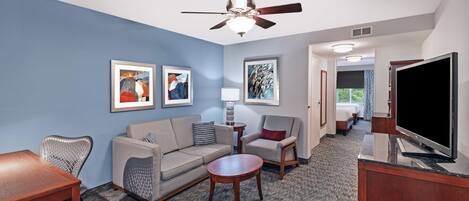 A hotel room with a blue accent wall, a ceiling fan, a flat-screen TV, a sofa, a coffee table, and a desk with a chair.