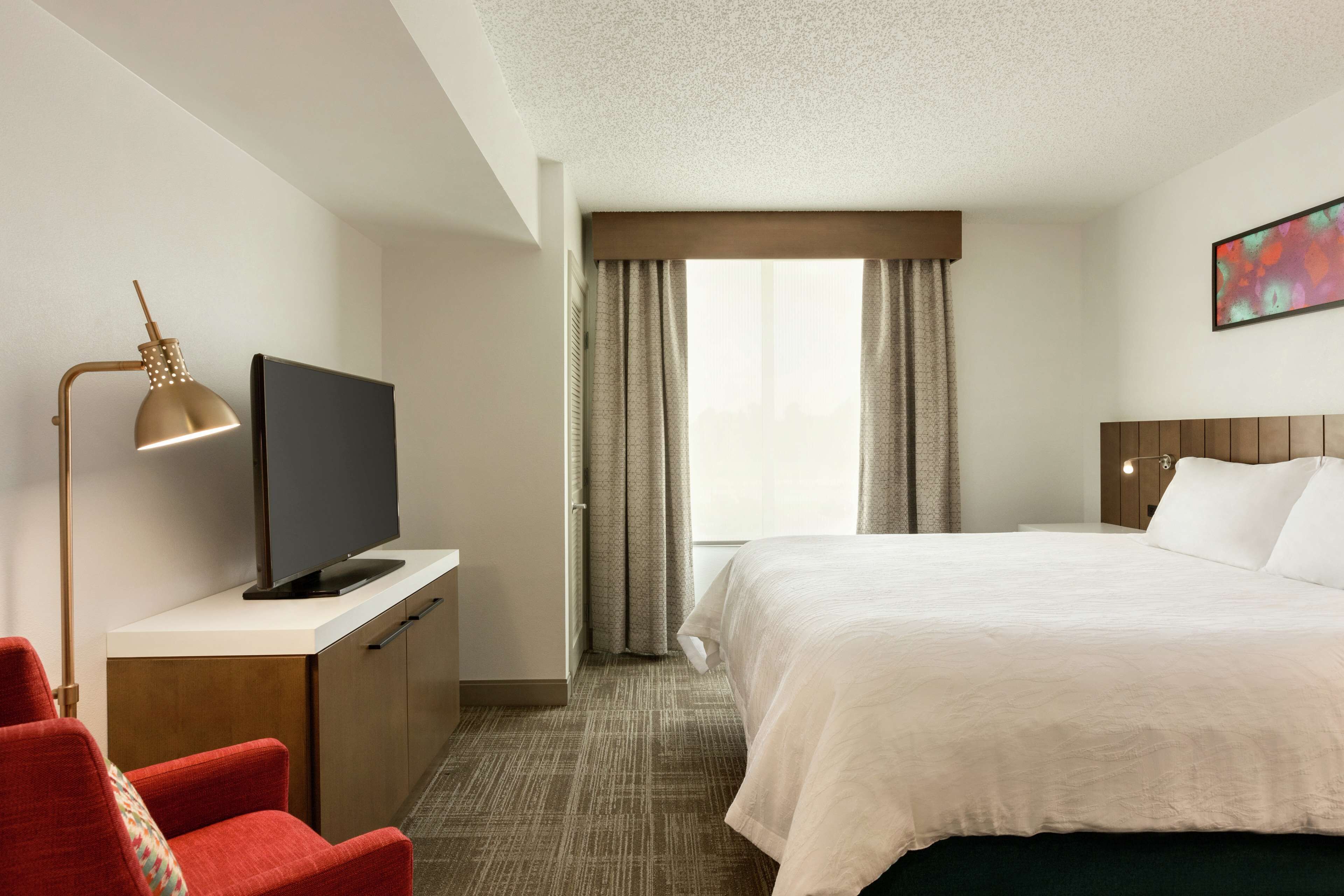 Quarto de hotel com cama, televisão em uma escrivaninha, uma cadeira vermelha e janela com cortinas.