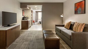 A modern hotel room with a flat-screen TV, a sofa, a bench, and a small kitchenette. - Hilton Garden Inn Tifton (Tifton)