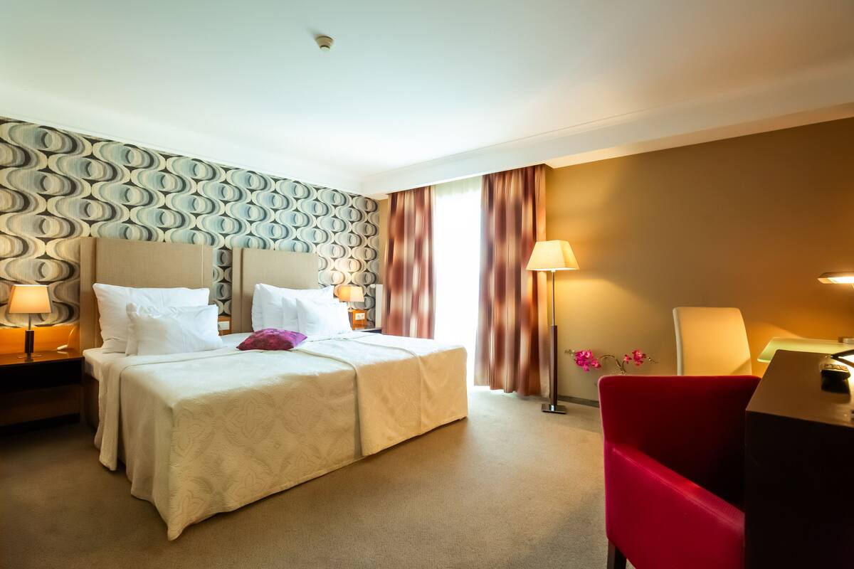 A hotel room with a bed, two bedside tables with lamps, a red chair, and a desk with a lamp.