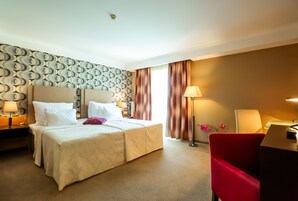 Una habitación de hotel con cama, dos mesitas de noche con lámparas, una silla roja y un escritorio con lámpara.