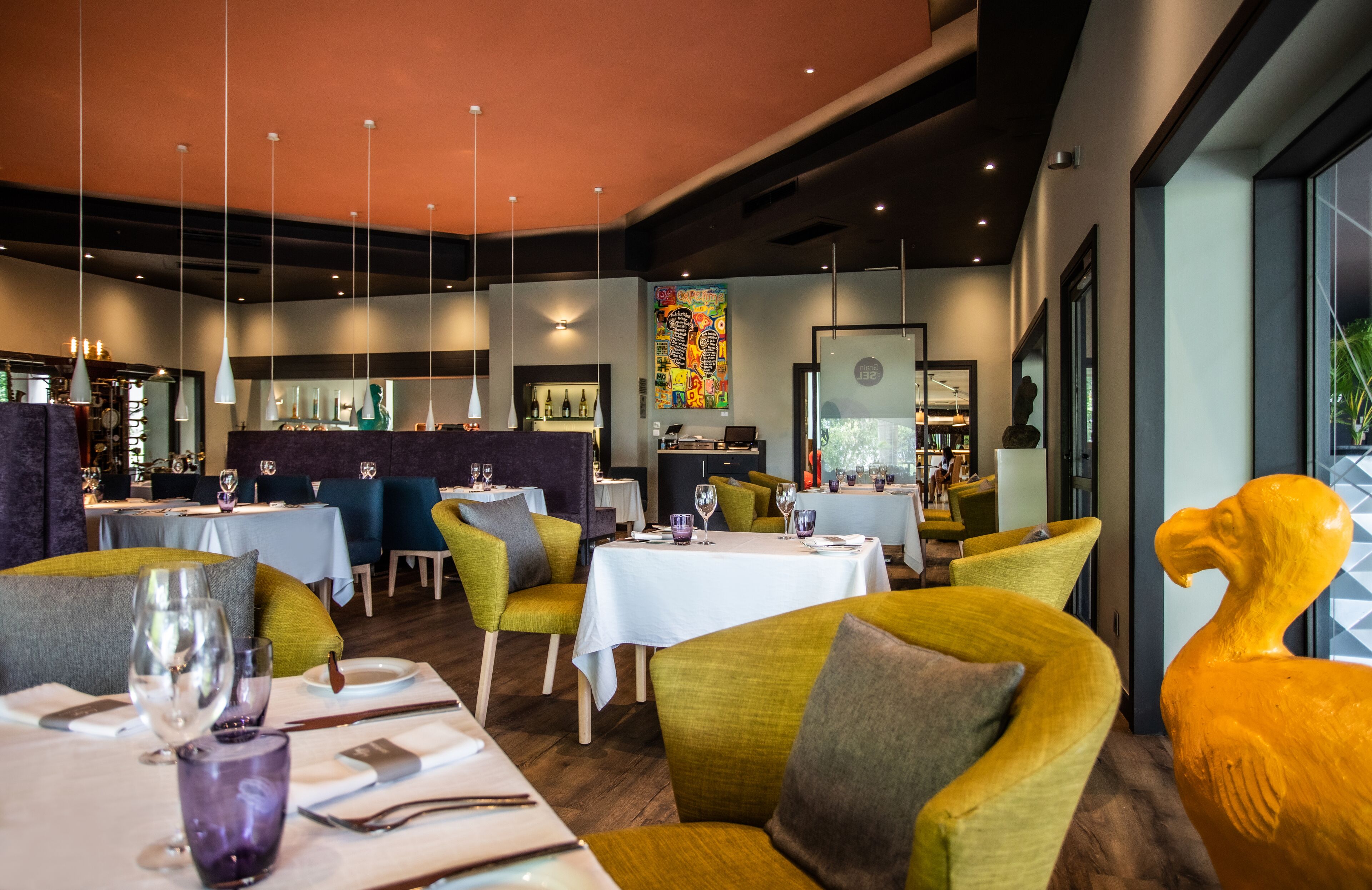 Restaurante moderno com pé-direito alto, iluminação pendente e cadeiras estofadas em tons de verde e amarelo. Mesas montadas com copos de vinho e talheres.