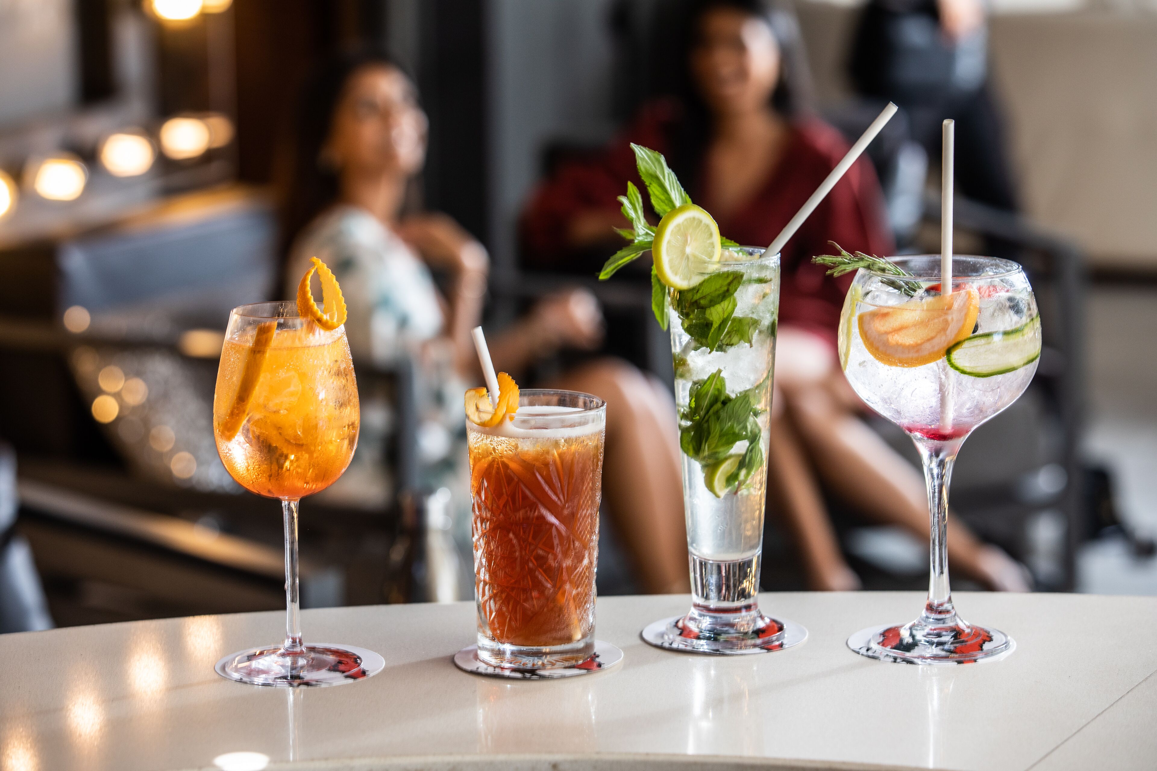 Four cocktails on a bar counter with a blurred background of people.