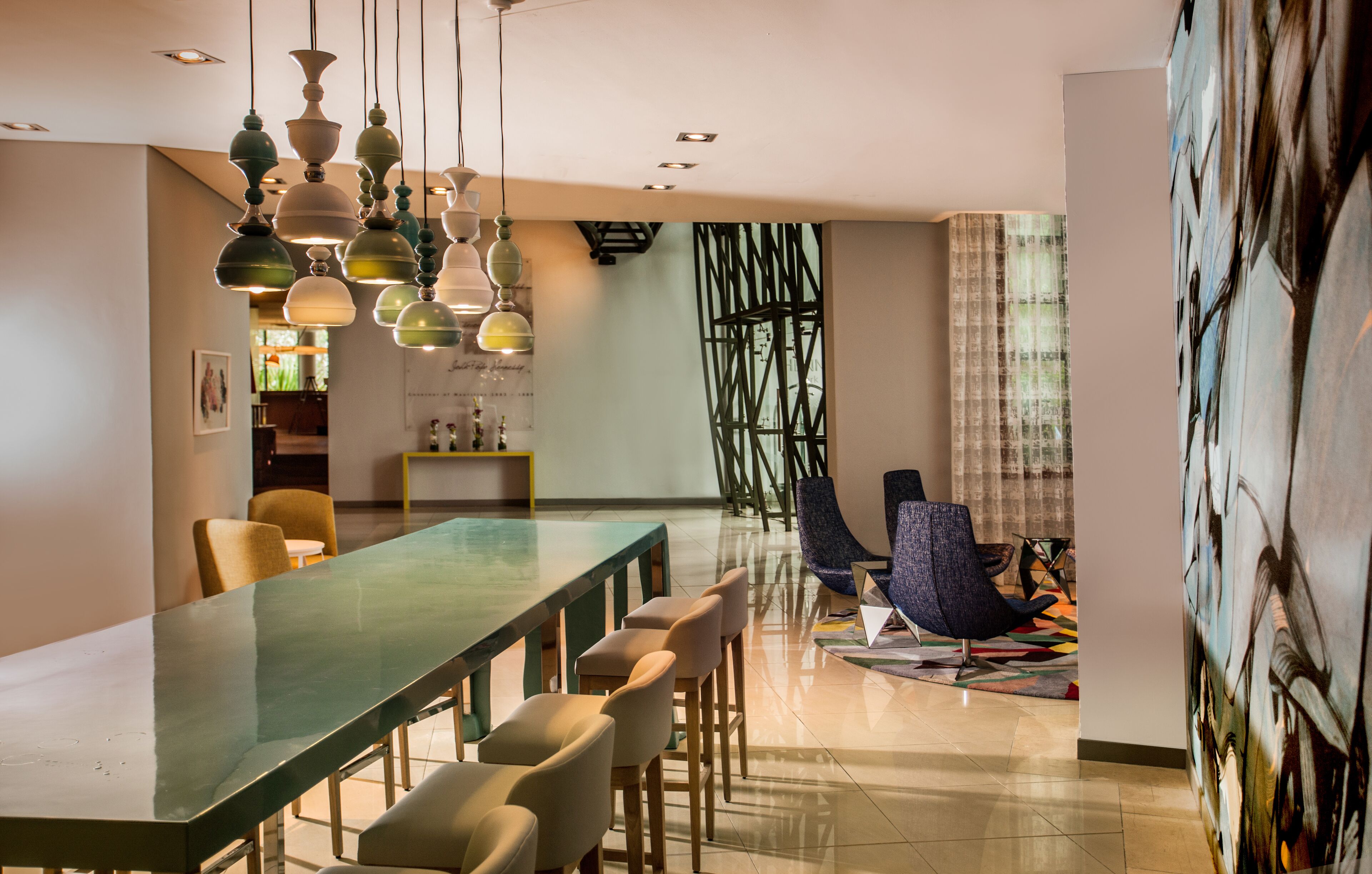 A modern dining area with a long table, multiple chairs, hanging pendant lights, and a contemporary art piece on the wall.