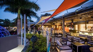 An outdoor seating area with tables and chairs, a bar area, and a palm tree. - Hennessy Park Hotel (Ebene)