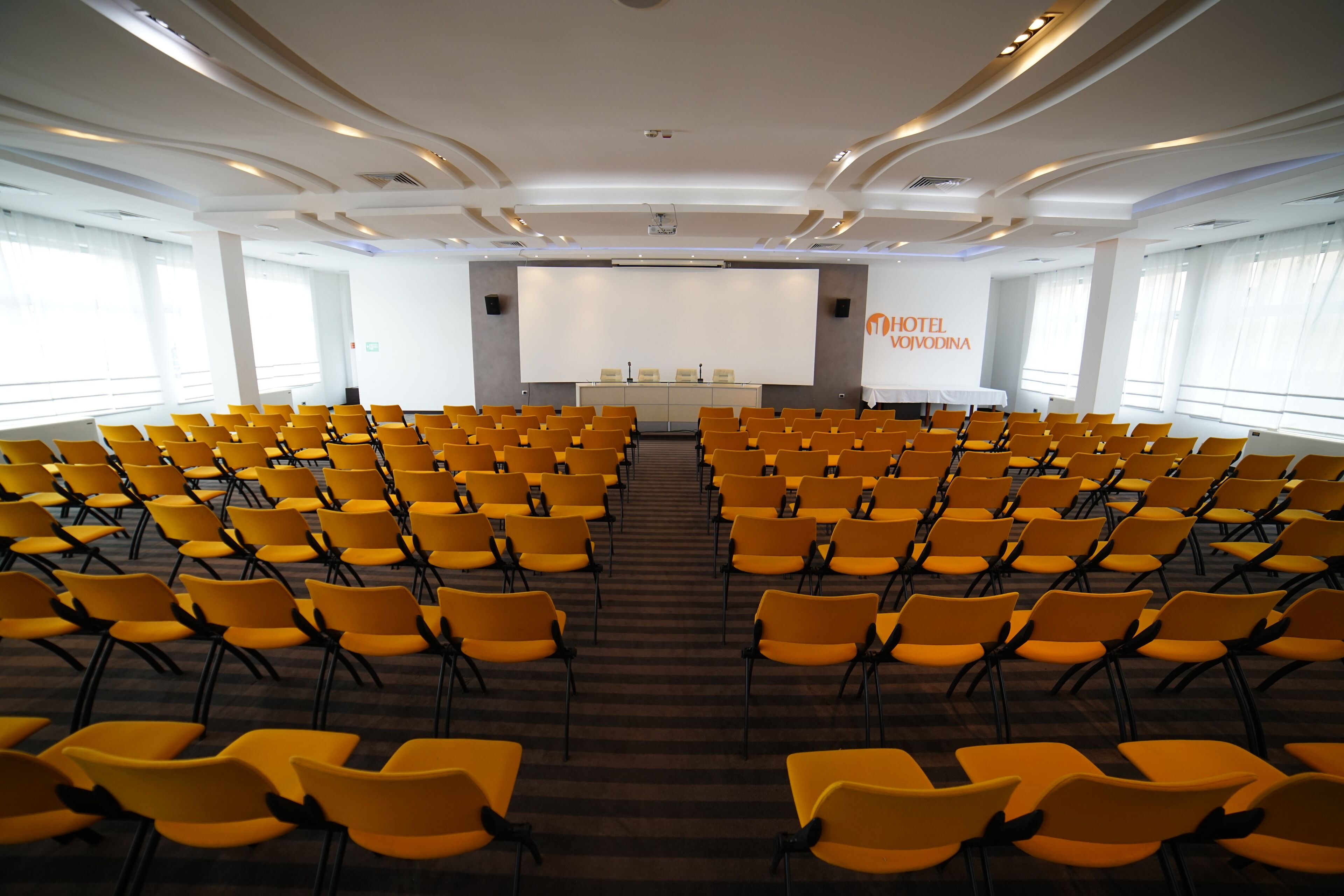 A conference room with rows of yellow chairs, a podium, and a large screen.