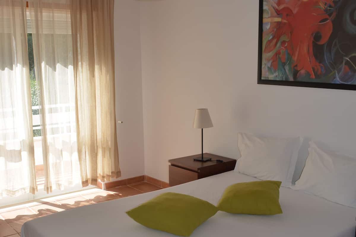 Habitación de hotel con una cama grande, una mesita de noche con lámpara, un cuadro en la pared y vistas al exterior a través de cortinas translúcidas.