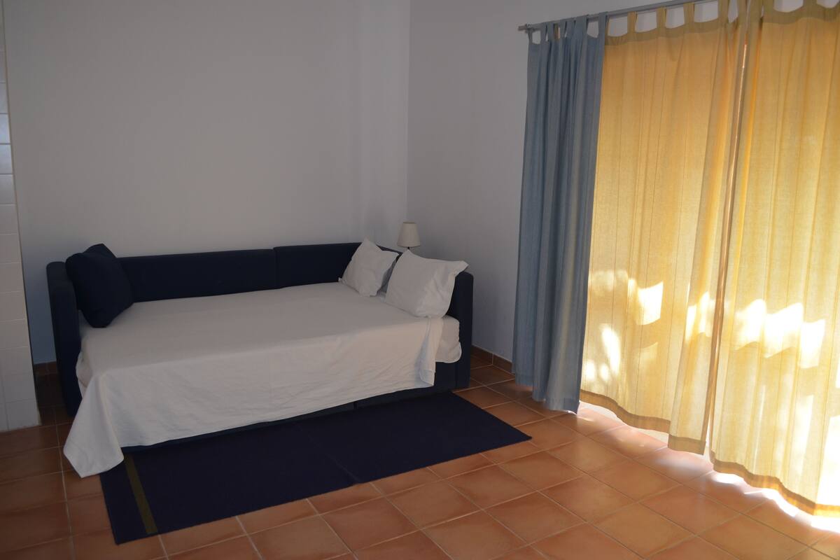 Una cama individual con sábana blanca en una habitación con piso de baldosas y ventana con cortinas amarillas.