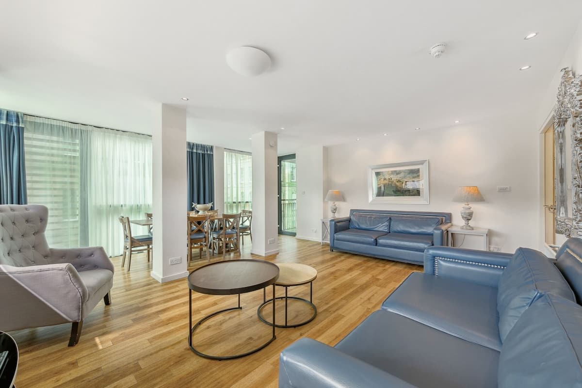 Una sala de estar moderna con un sofá azul, un sillón gris y una mesa de centro redonda. Se observa una zona de comedor con una mesa y sillas al fondo.