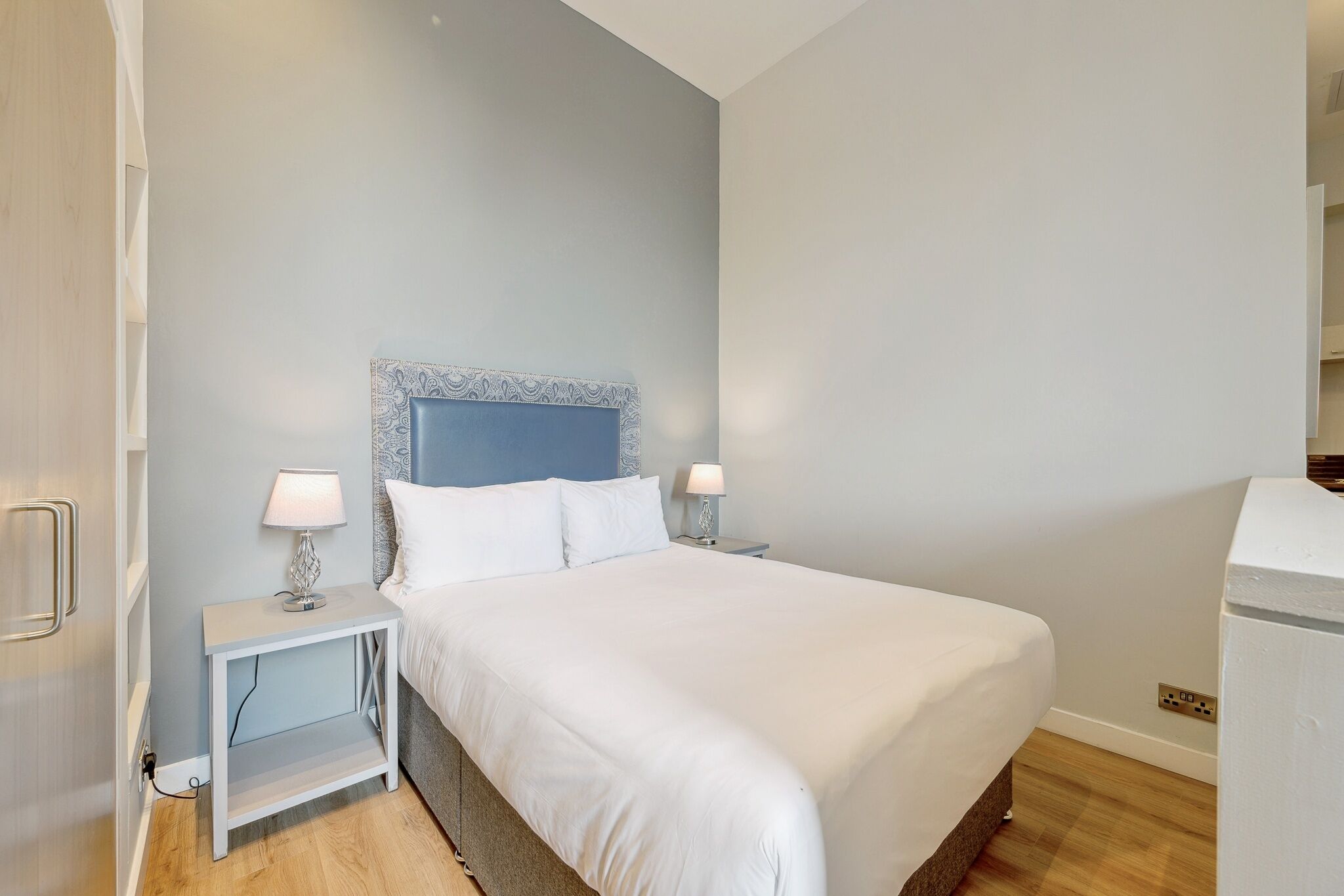 A modern hotel room with a large bed, two bedside tables with lamps, a blue headboard, and a white wardrobe.