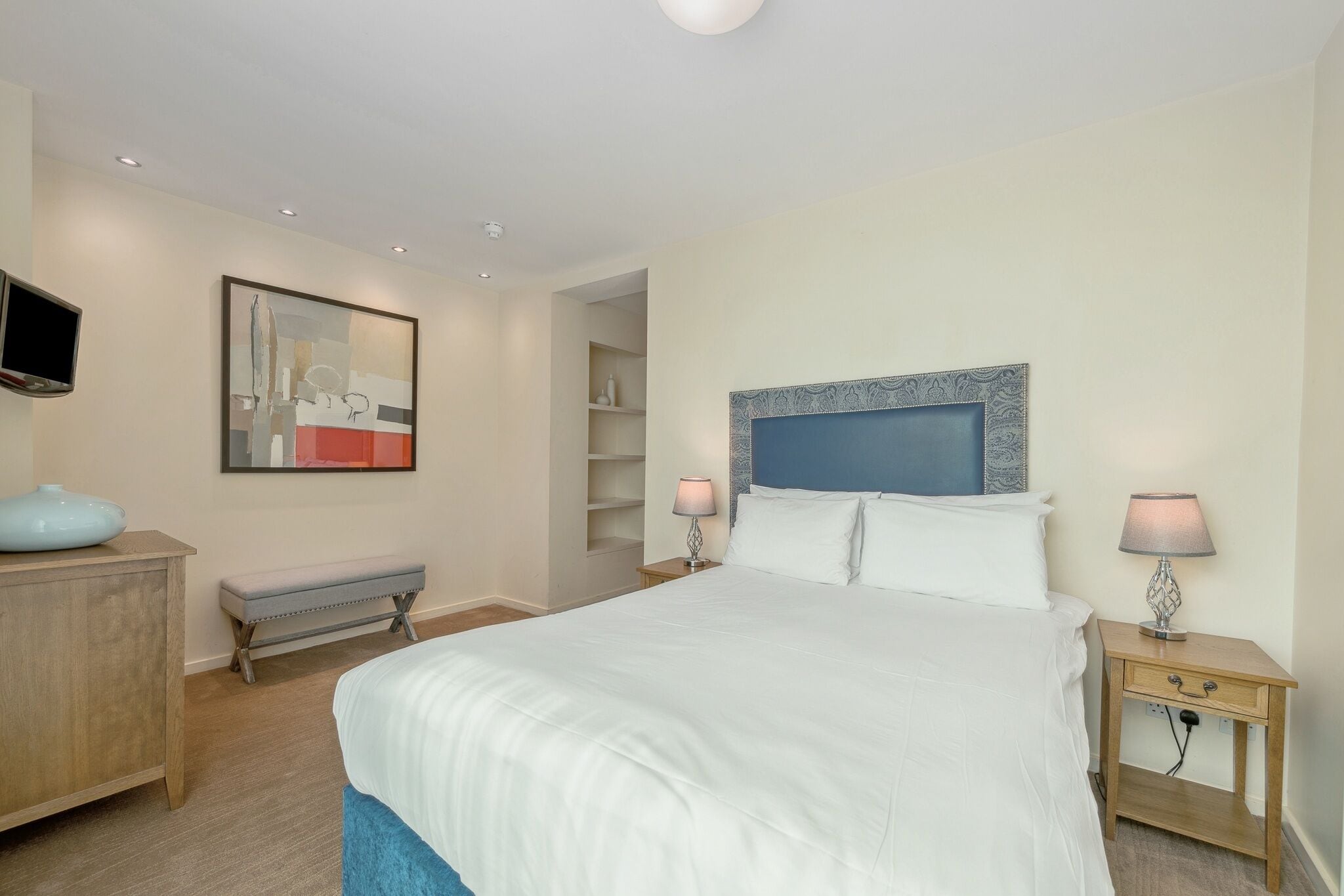 Een slaapkamer met een groot bed, nachtkastjes, een bankje, een televisie en kunstwerken aan de muur.