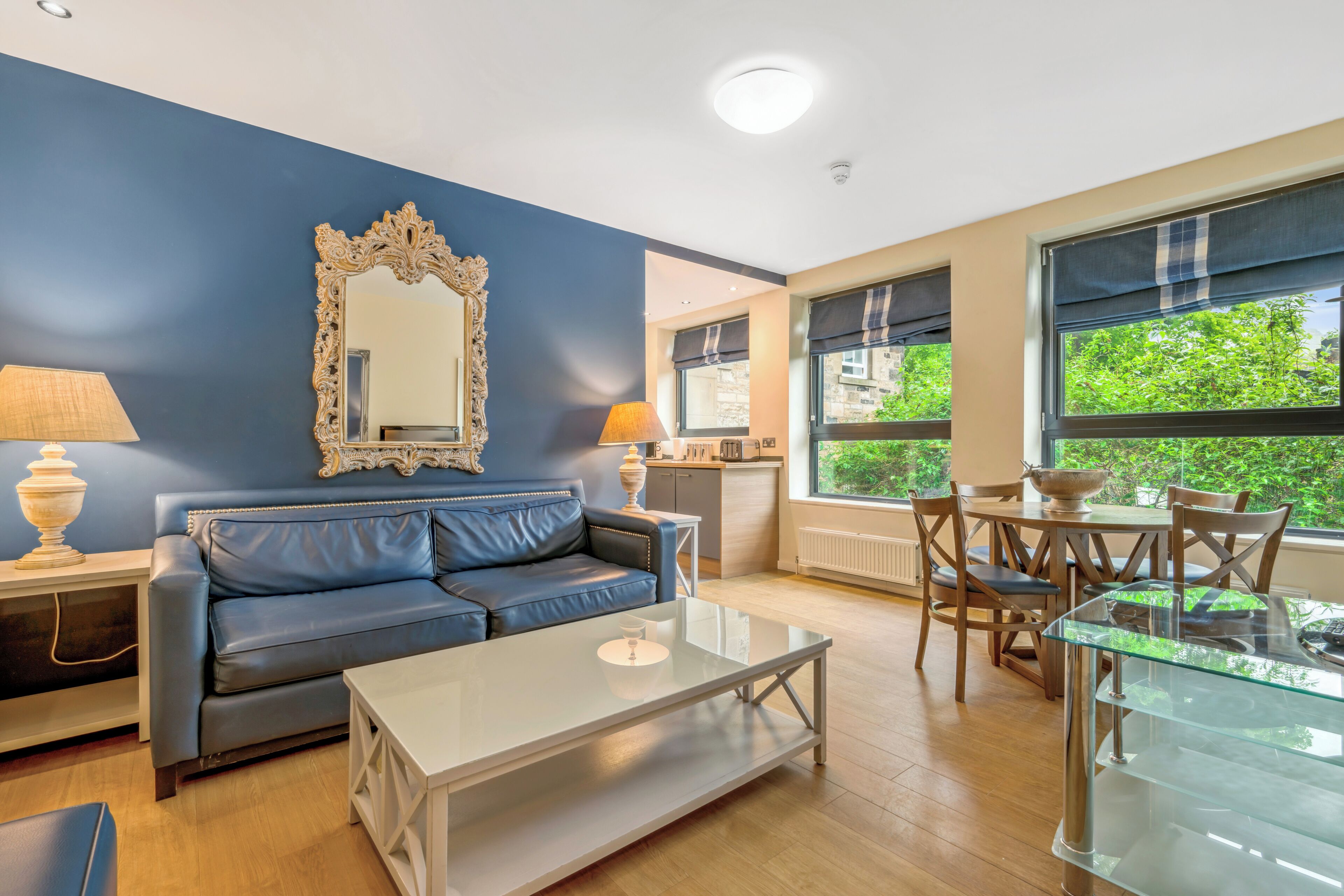 Ein modernes Wohnzimmer mit einem blauen Sofa, einem Glastisch, einem Zier-Spiegel und einem Essbereich mit Tisch und Stühlen.