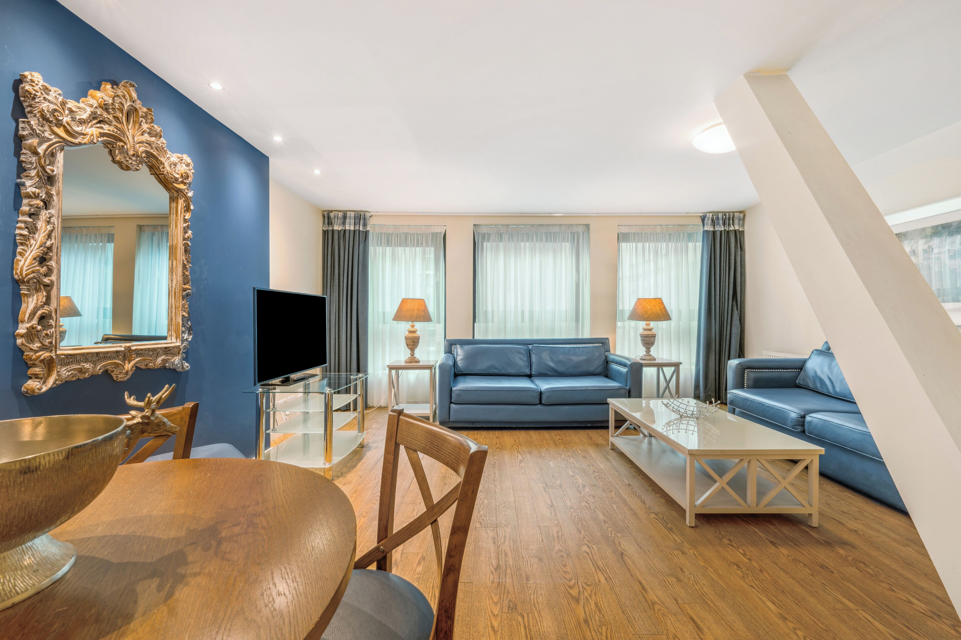 A modern living room with a blue sofa, a television, and a dining area with a wooden table and chairs.