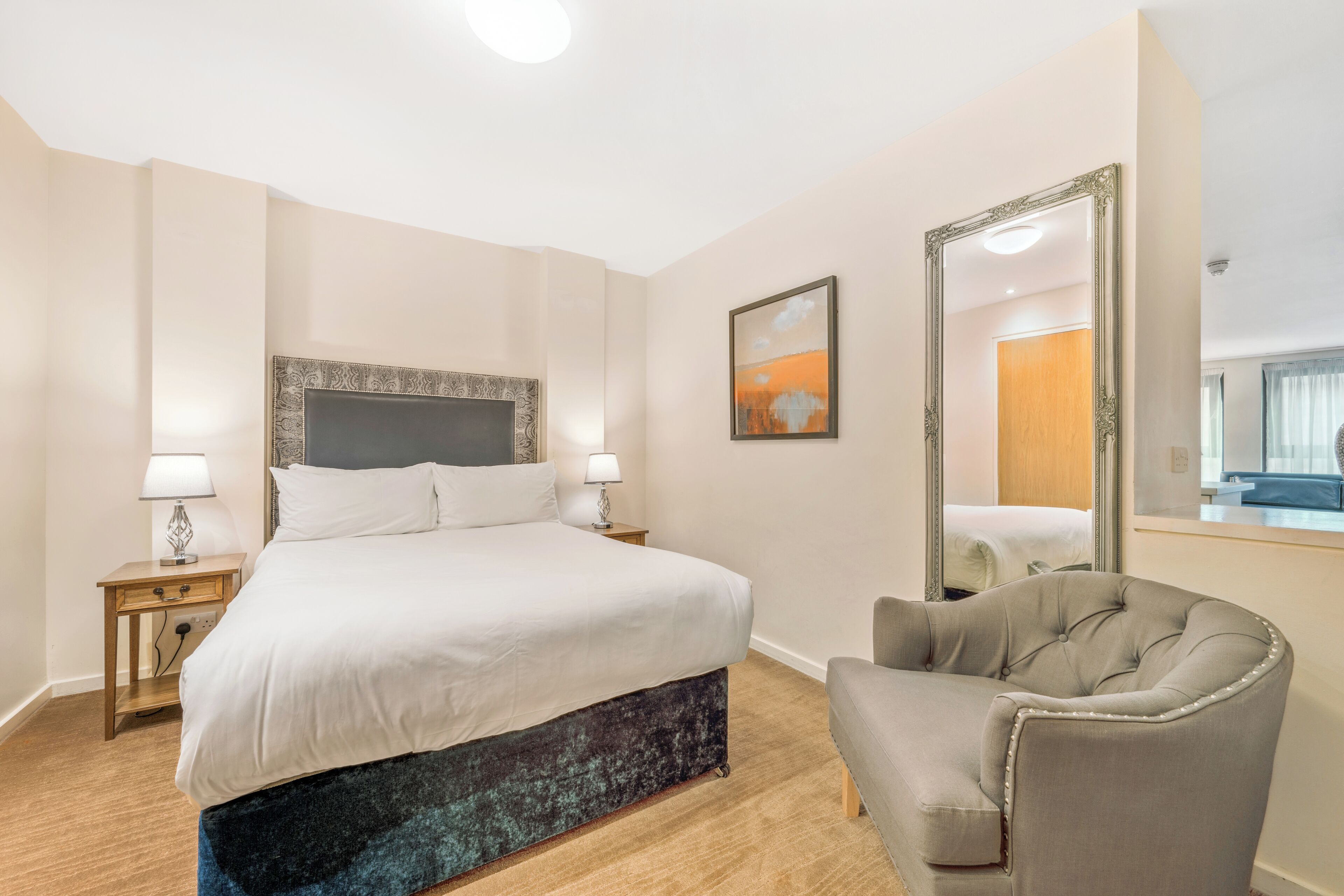 Une chambre d’hôtel avec un grand lit, des tables de chevet avec des lampes, une chaise, un grand miroir et un tableau au mur.