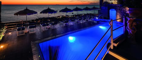 Zona junto a la piscina con sillones reclinables y sombrillas, con vistas al mar al atardecer.