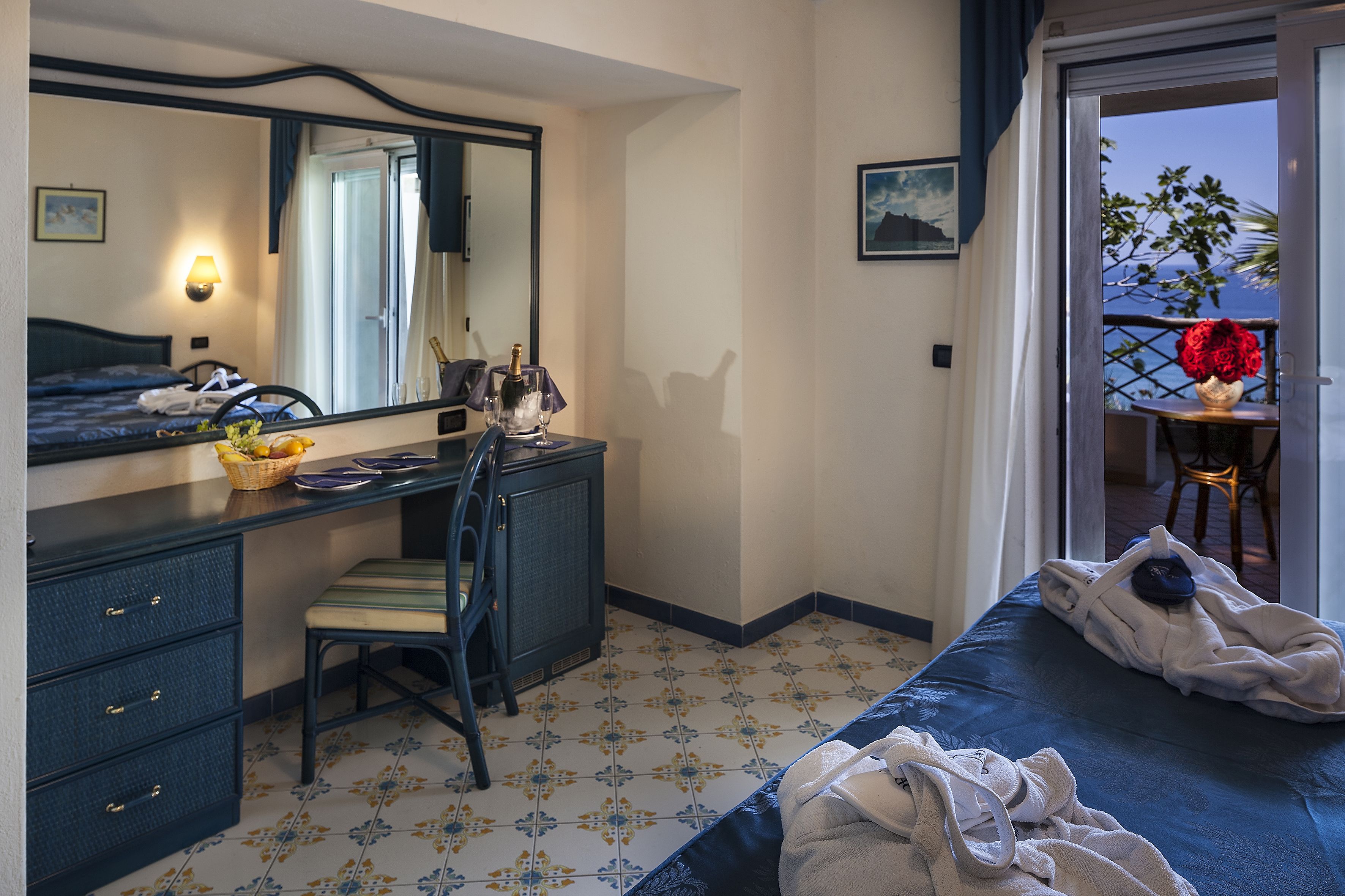 A hotel room with a bed, a desk with a chair, a dresser, a mirror, and a balcony with a table and a vase of flowers.