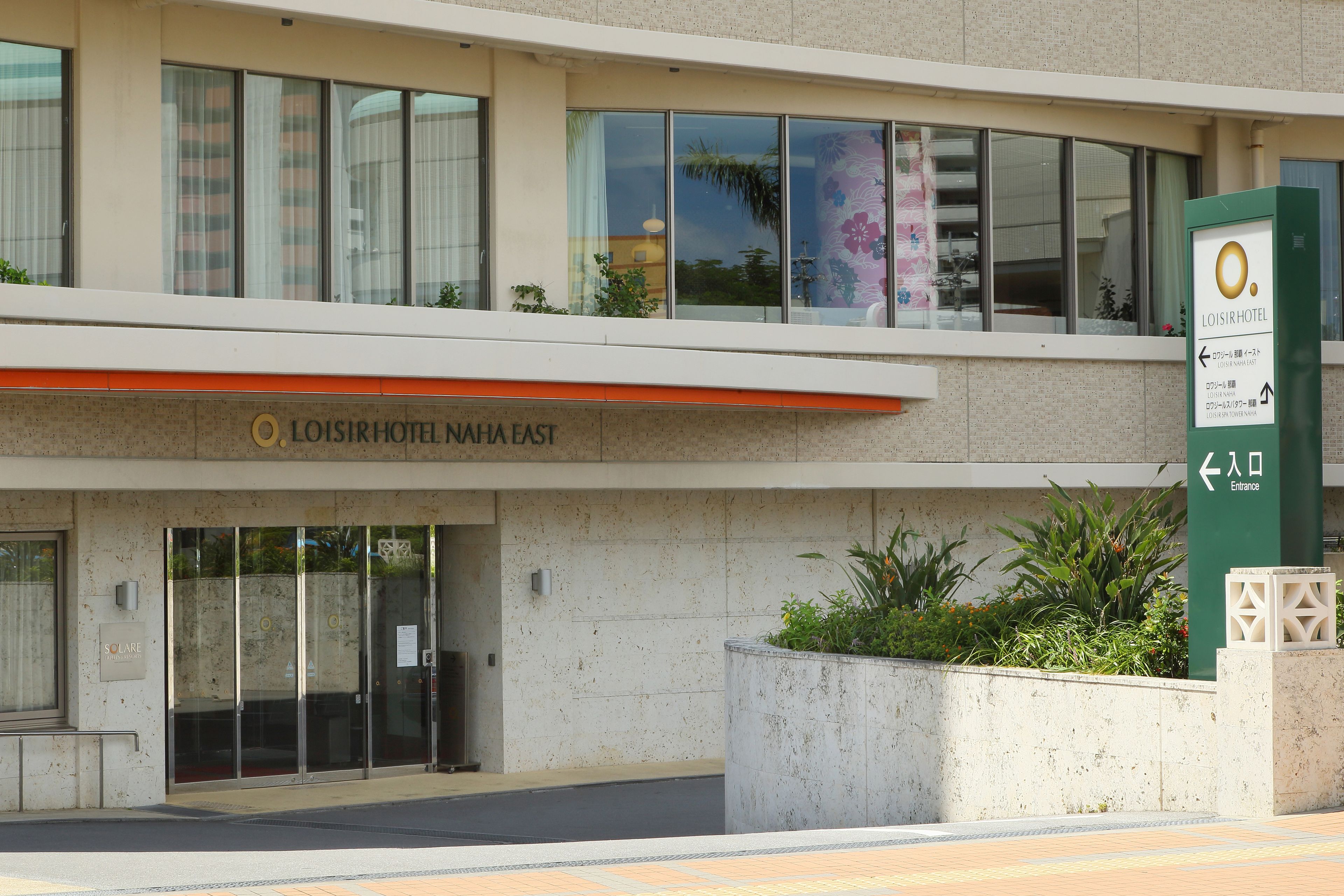 The exterior of Loisir Hotel Naha East with a clear entrance, a signboard, and a landscaped area with plants.