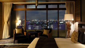 Habitación de hotel con un amplio ventanal que ofrece vistas a la ciudad, una cama, dos sillones, una mesita y una lámpara.