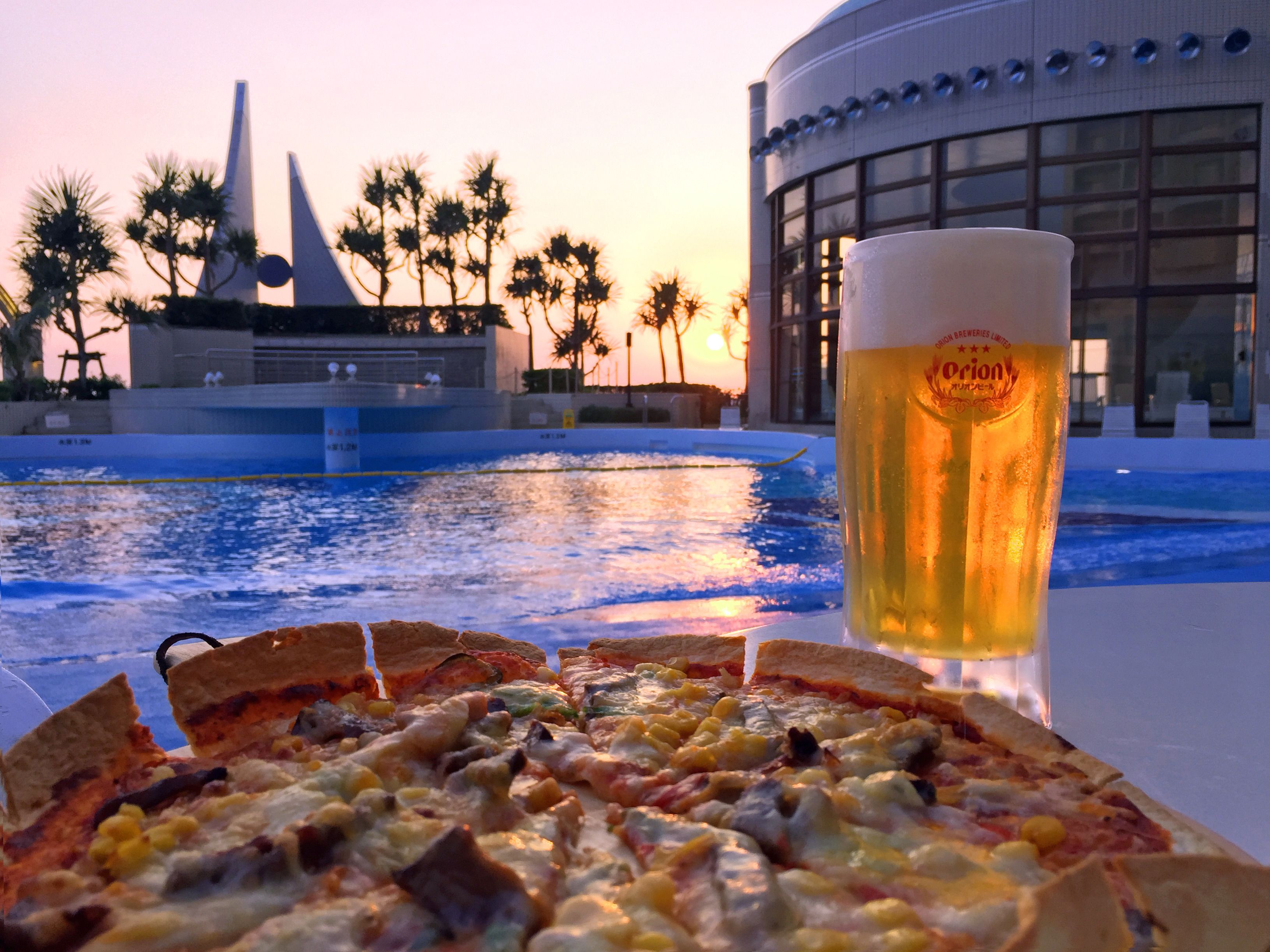 Pizza con diferentes ingredientes y una cerveza en una mesa junto a una piscina, con palmeras y un edificio al fondo.