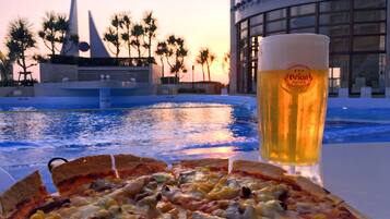 Pizza con diferentes ingredientes y una cerveza en una mesa junto a una piscina, con palmeras y un edificio al fondo.