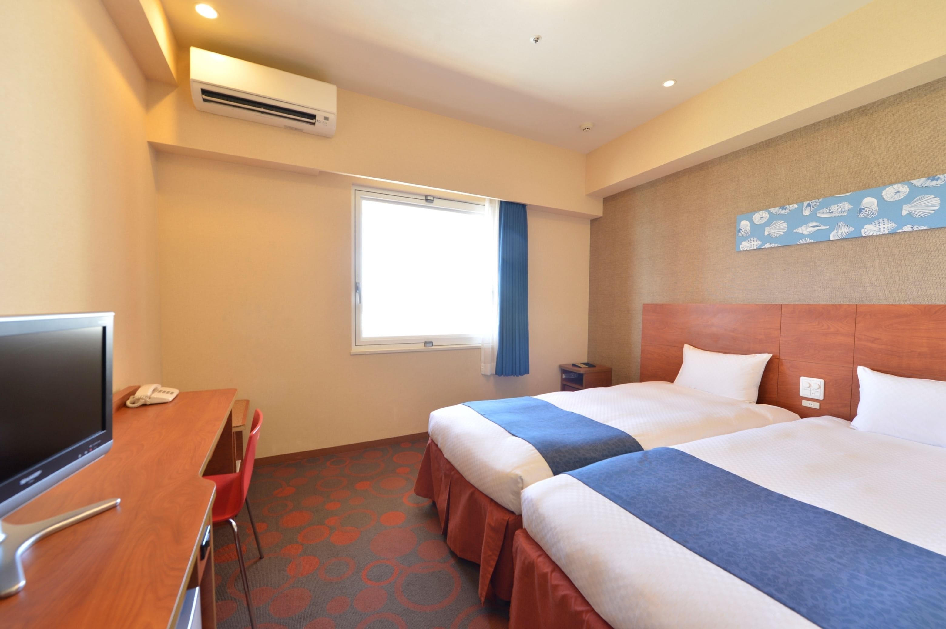 A hotel room with two beds, a desk with a TV, and a window with curtains.