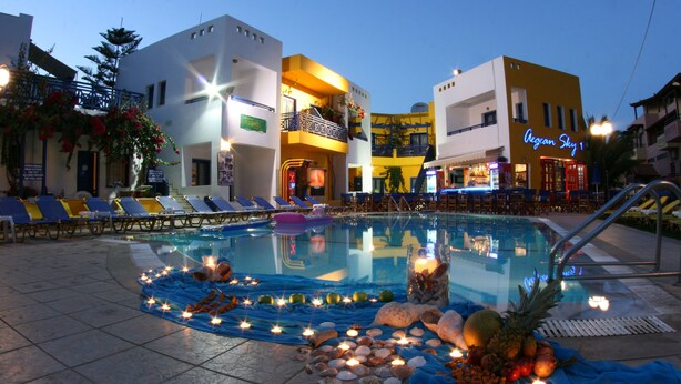 A pool area with lounge chairs, a swimming pool, and a building with a neon sign. - Aegean Sky Hotel & Suites (Hersonissos)