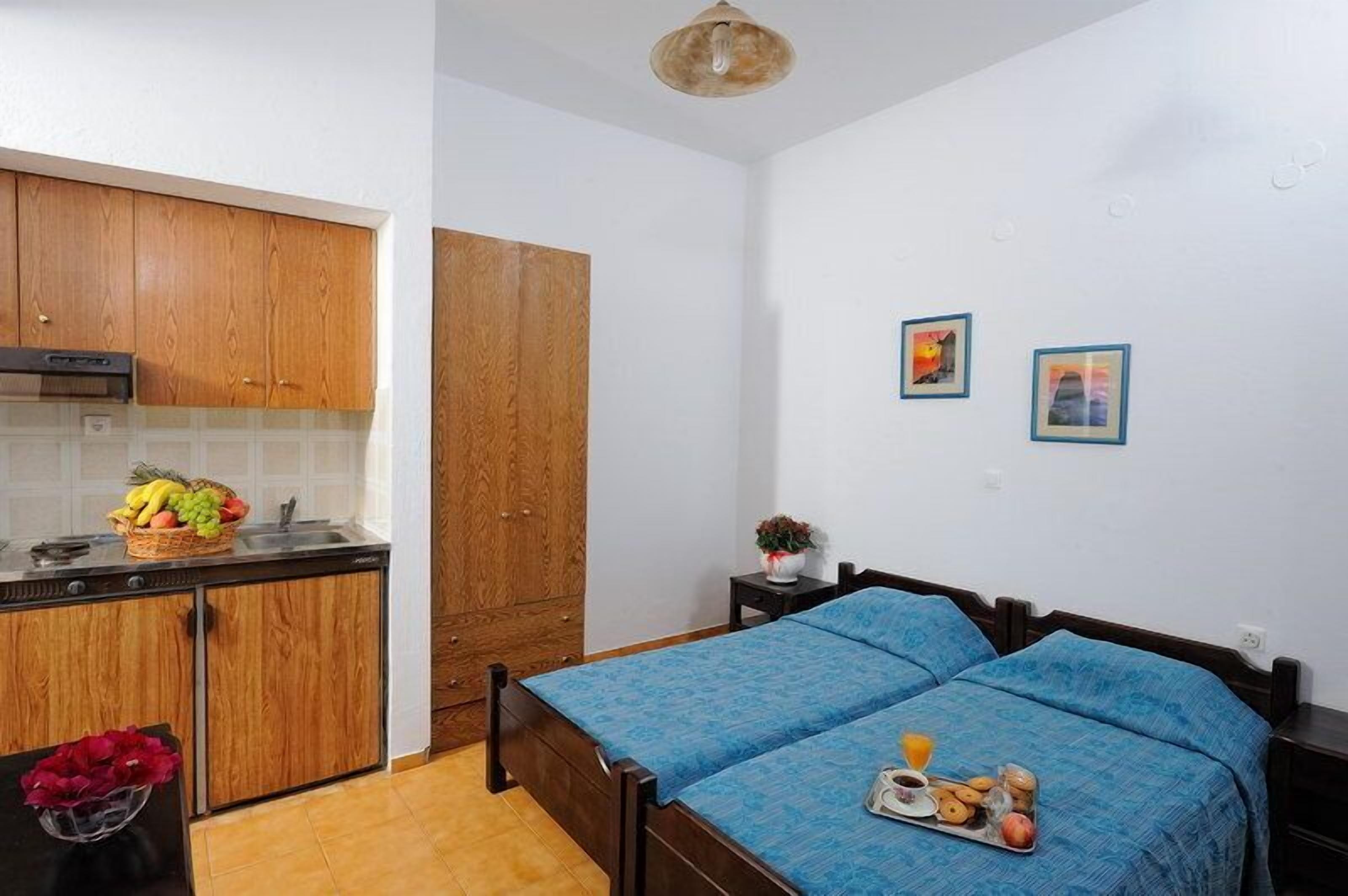 A bedroom with a bed, a bedside table, and a nightstand. There is a kitchen area with wooden cabinets and a countertop.