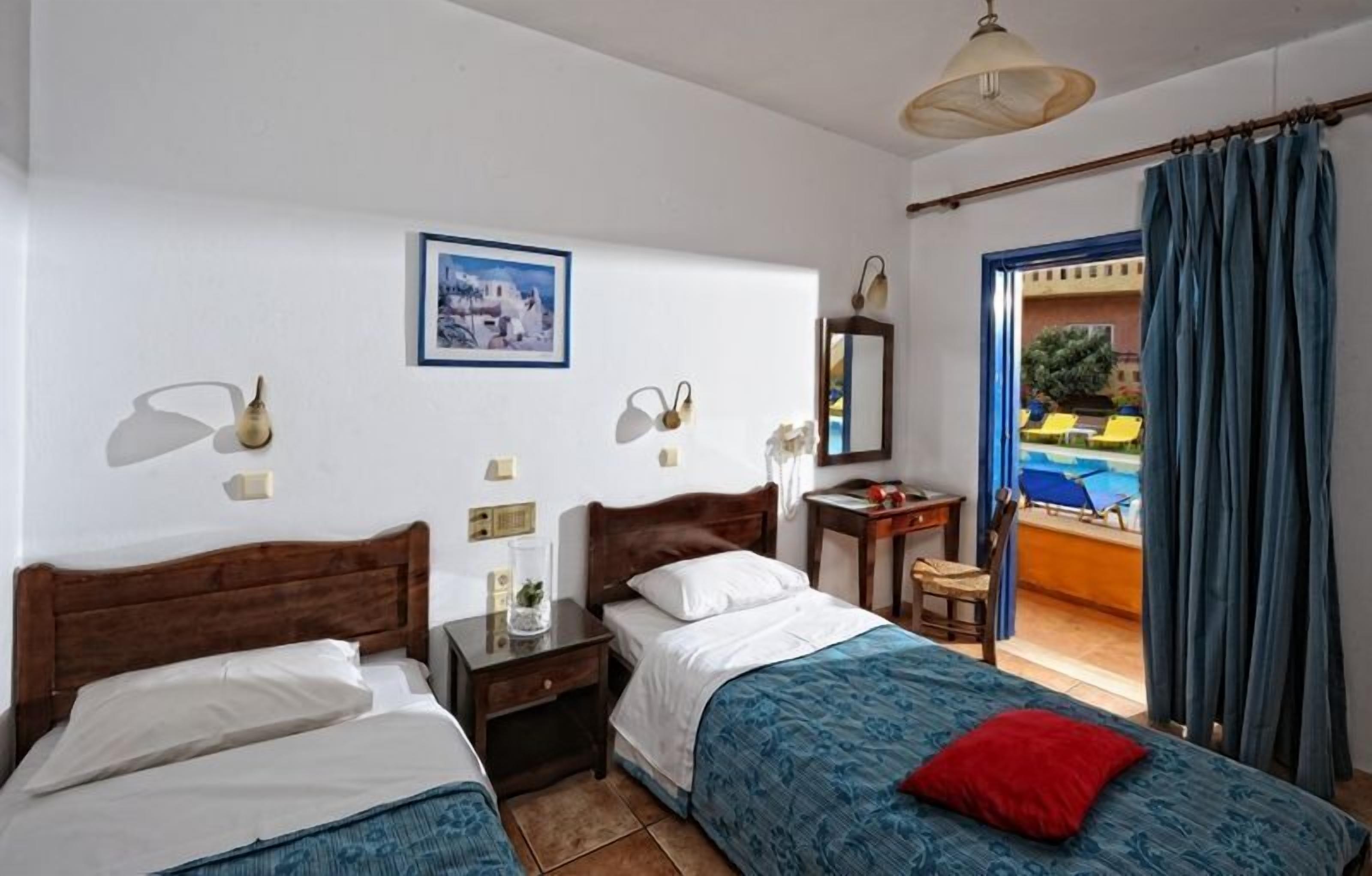 A hotel room with two beds, a small table, a nightstand, a framed picture, and a view of a pool through a door.