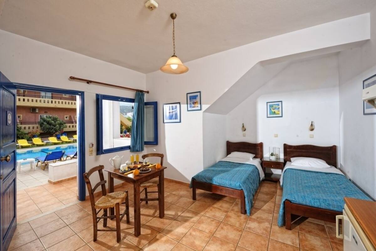 A bedroom with two beds, a dining table with chairs, a window with curtains, and a view of a swimming pool outside.