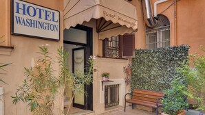 A hotel entrance with a sign reading "HOTEL WASHINGTON," a bench, potted plants, and a decorative wall.