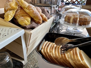 Una exhibición de diversos productos de panadería, incluyendo croissants y pan, con una bandeja de pan en rebanadas y una cuchara.