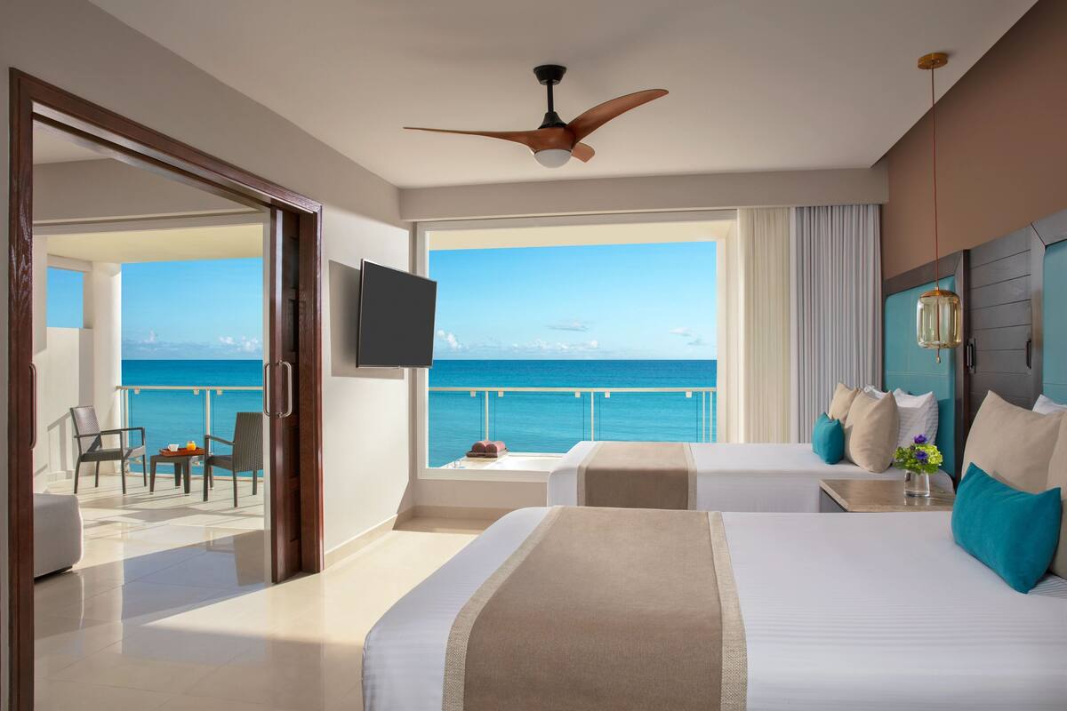 A hotel room with two beds, a ceiling fan, a TV, and a balcony with a view of the ocean.