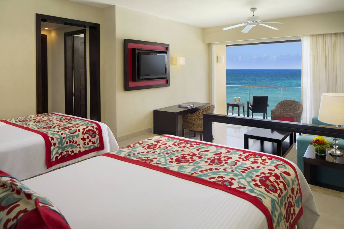 A hotel room with two beds, a desk, a chair, a TV, and a balcony with a view of the ocean.