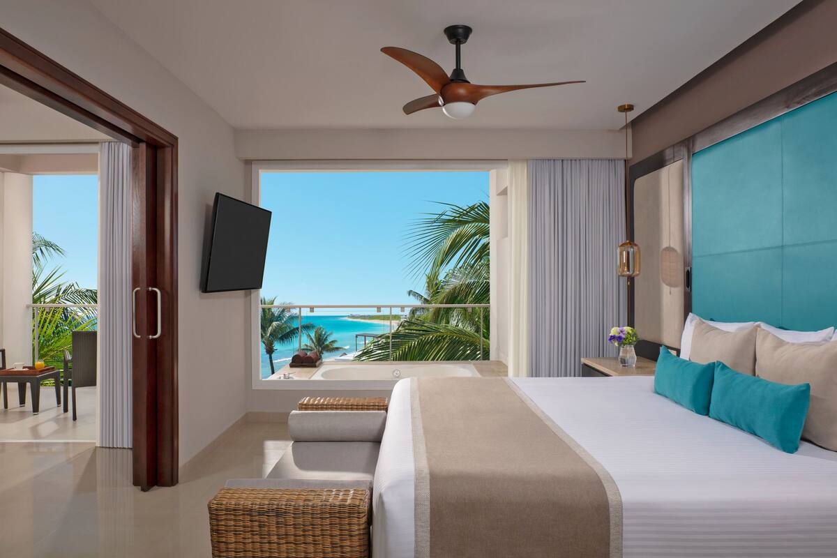 A hotel room with a bed, a TV, a ceiling fan, a balcony with a view of the ocean, and a dining area.