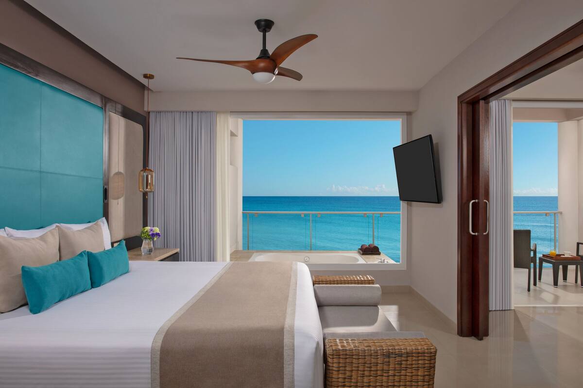 A modern hotel room with a large bed, a ceiling fan, a TV, and a balcony with a view of the ocean.