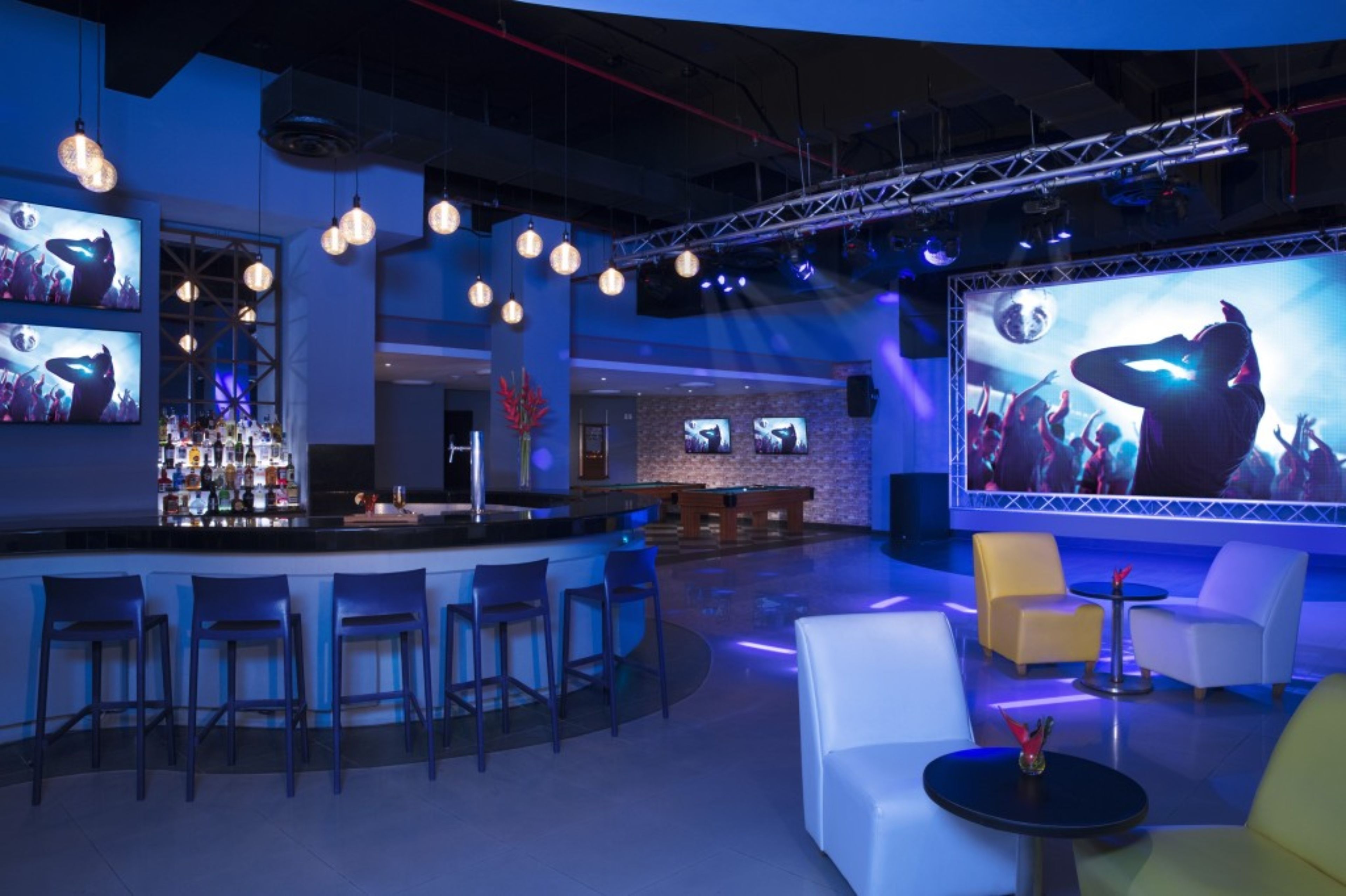 A bar area with multiple stools, a bar counter, and a large screen displaying a concert scene.