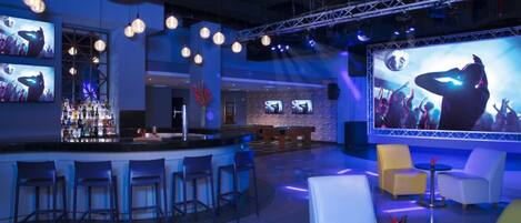 A bar area with multiple stools, a bar counter, and a large screen displaying a concert scene.